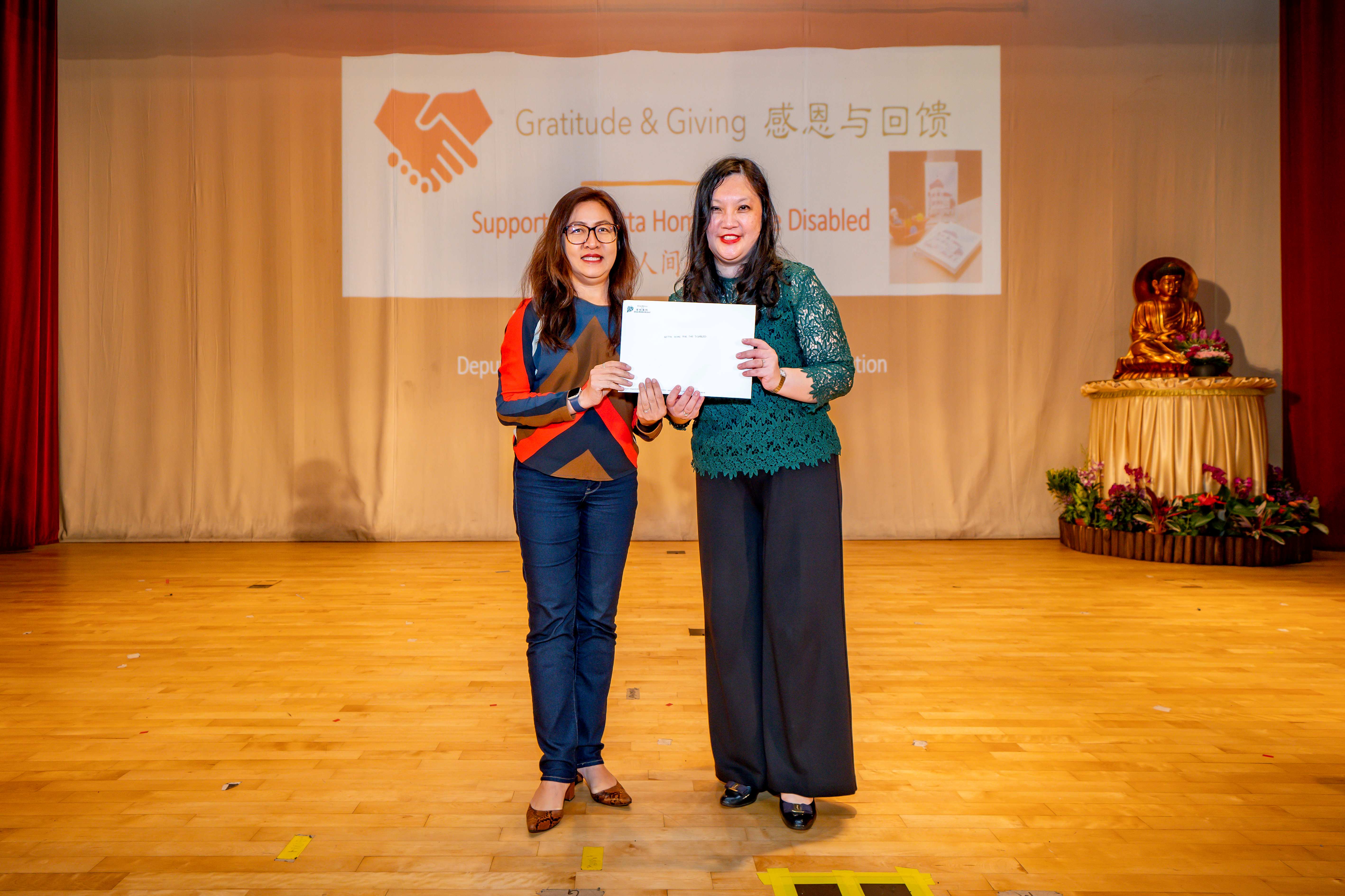 Two women stand on a wooden stage holding a certificate before a Gratitude and Giving event banner.
